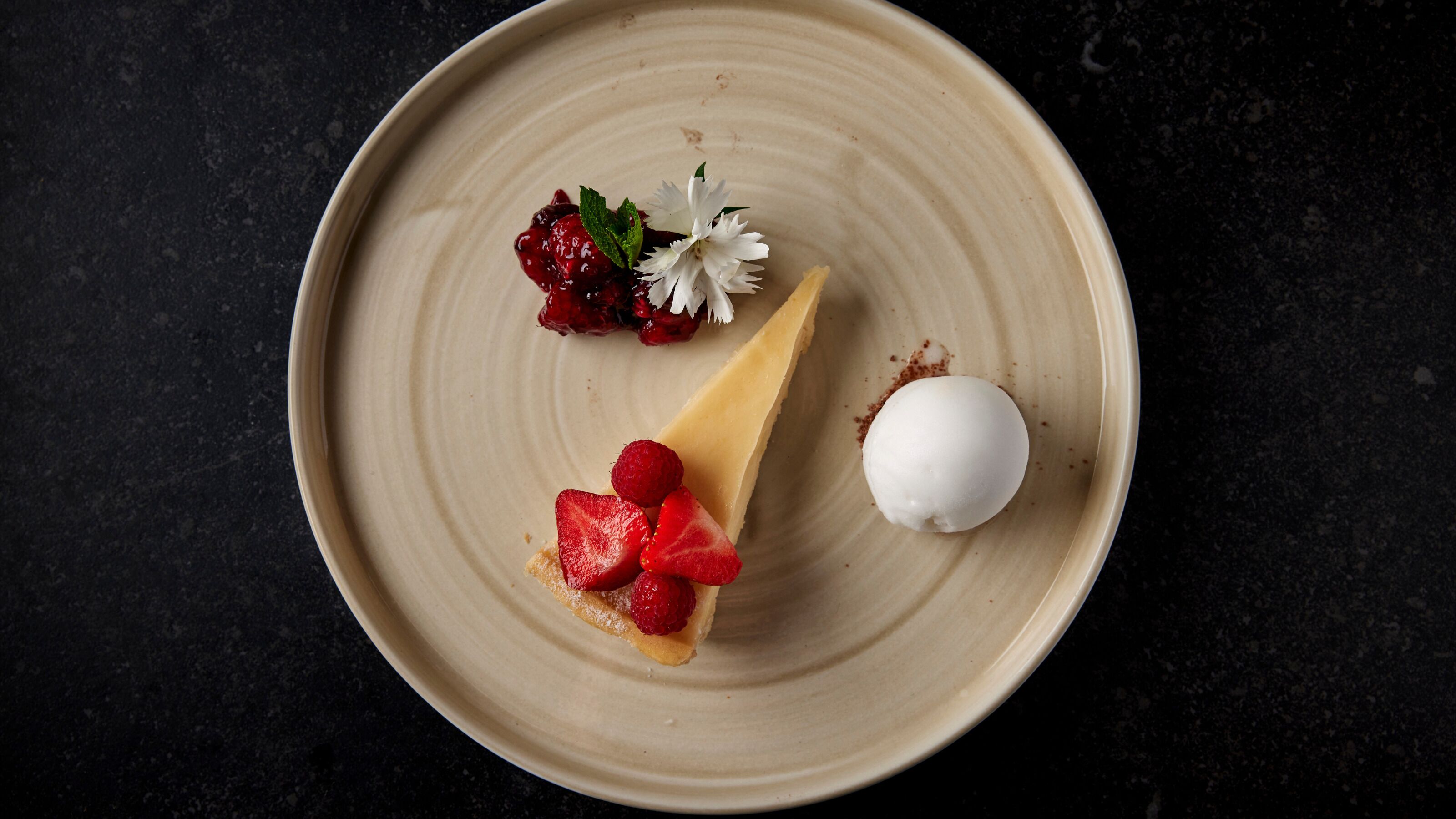 A dessert plate with a slice of cheesecake topped with strawberries and raspberries, a scoop of vanilla ice cream, and a small white flower.