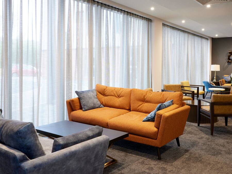 A modern hotel room with a large window, a sofa, a coffee table, and a dining area with chairs and a table.