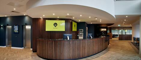 A hotel reception area with a curved reception desk, wooden paneling, and a herringbone wood floor. There are signs for 'Newport' and 'JASA' on the wall, and a reception desk with computers.