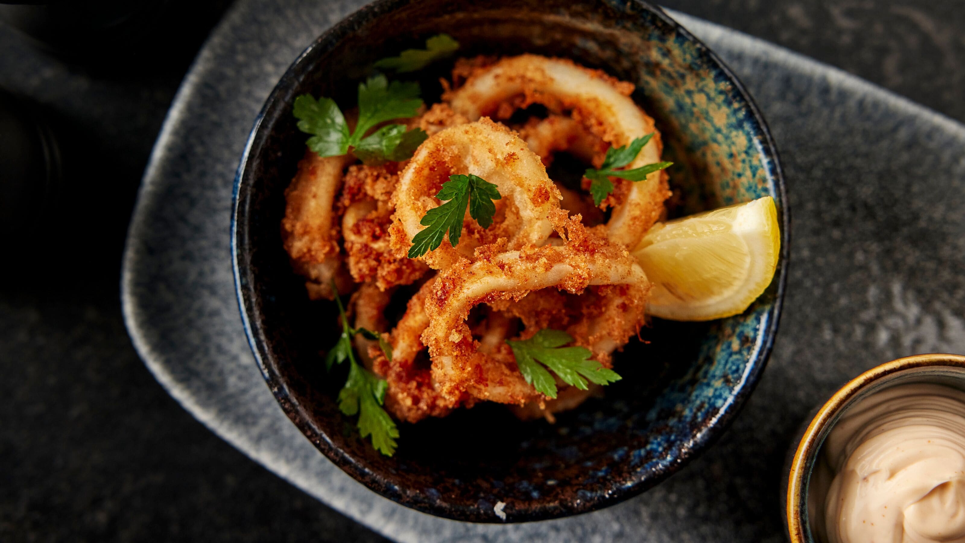 A bowl of fried calamari garnished with parsley and a lemon wedge, placed on a dark surface.