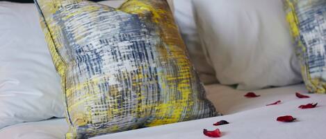 A bed with white and patterned pillows, some red petals scattered on the bed.
