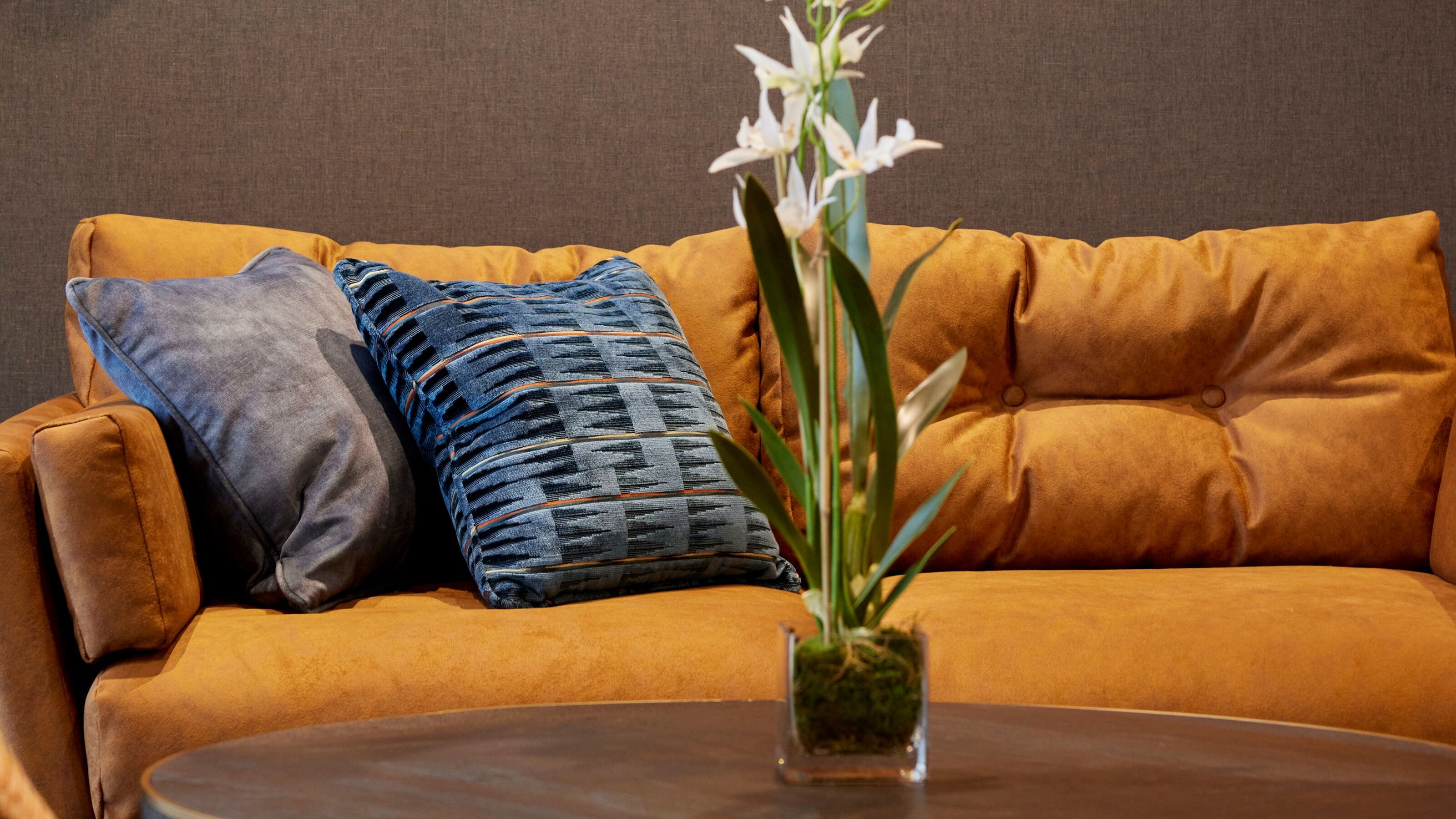 A sofa with two pillows, a small vase with a plant, and a round table.