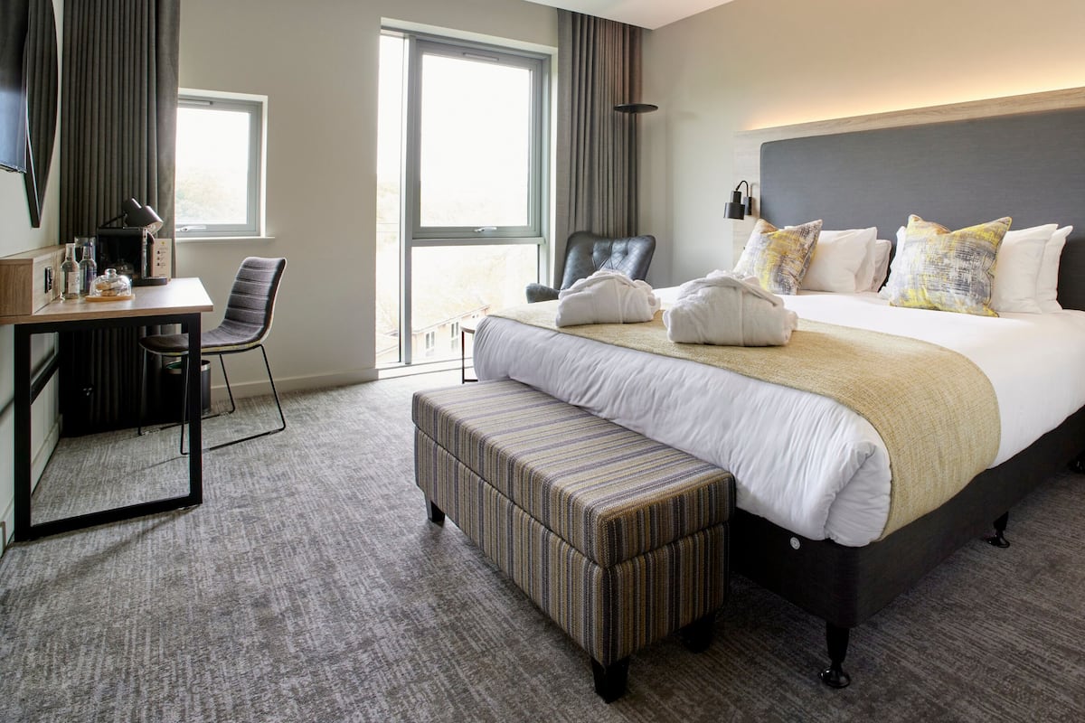 A hotel room with a large bed, a desk, a chair, and a bench.