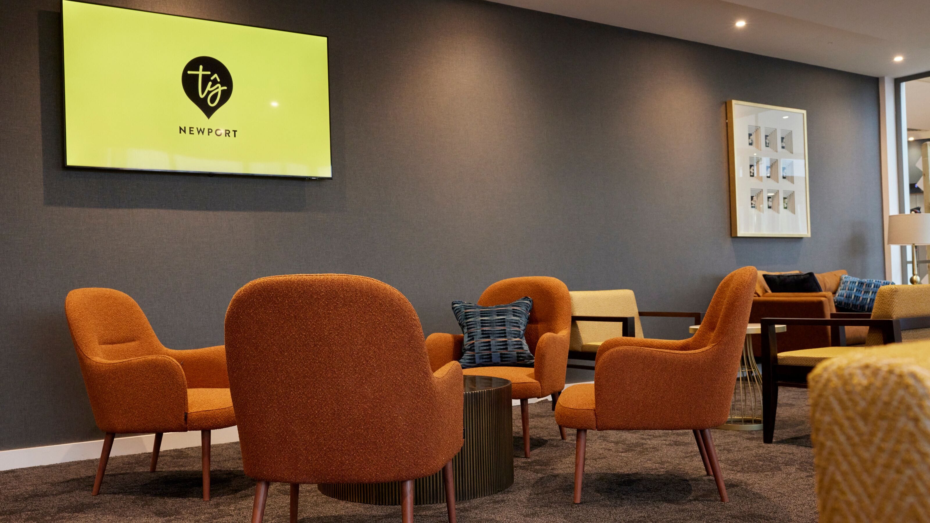 Un vestíbulo de hotel con sillones anaranjados, una mesa de centro y un televisor de pared mostrando el logotipo y el nombre 'NEWPORT'.