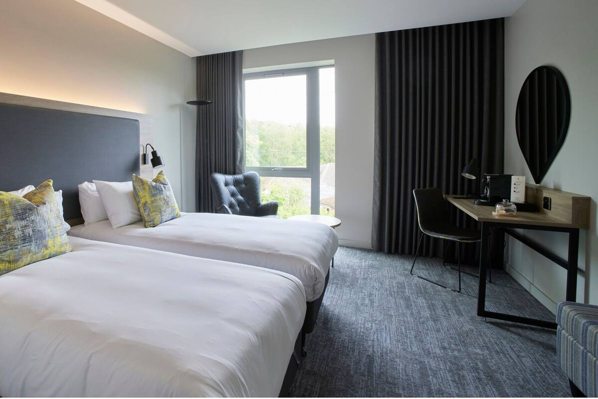 A hotel room with two beds, a desk, a chair, and a large window.