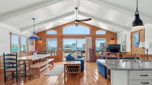 A spacious living area with a wooden ceiling, large windows, a blue sofa, and a TV. - In Due Tide: Oceanside, Hot Tub, Dogs OK (Rodanthe)
