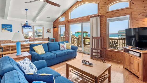 A spacious living area with a large sofa, a coffee table, and a television. There are wooden walls and large windows providing views of the outdoors. - In Due Tide: Oceanside, Hot Tub, Dogs OK (Rodanthe)
