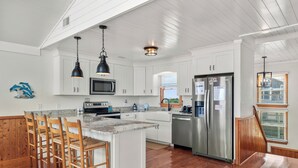 A spacious kitchen with white cabinetry, a stainless steel refrigerator, and a wooden island with bar stools. - In Due Tide: Oceanside, Hot Tub, Dogs OK (Rodanthe)