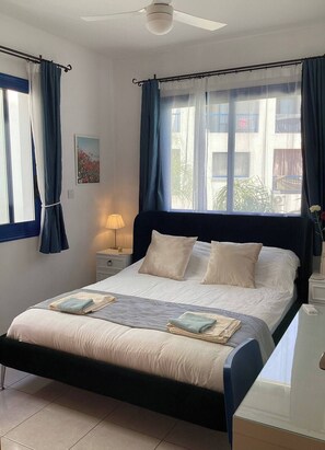A neatly made bed with a headboard, a bedside lamp, and a view of the outside through a window with curtains.
