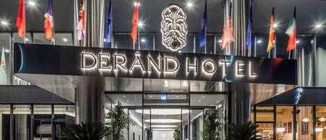 The entrance of Derand Hotel with a modern facade, a red carpet, and multiple flags.