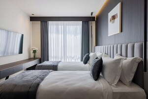 Quarto de hotel moderno com uma cama grande, mesa de cabeceira, TV de tela plana e uma janela ampla com cortinas.
