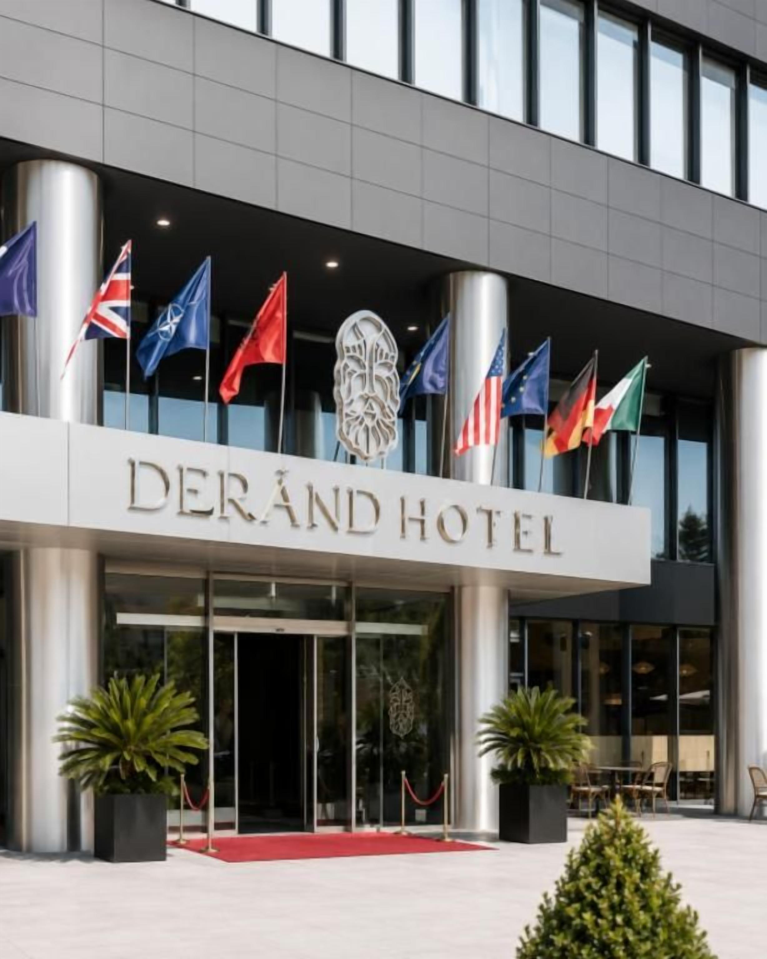 El exterior del Hotel Derand con varias banderas, una alfombra roja en la entrada y plantas en macetas.