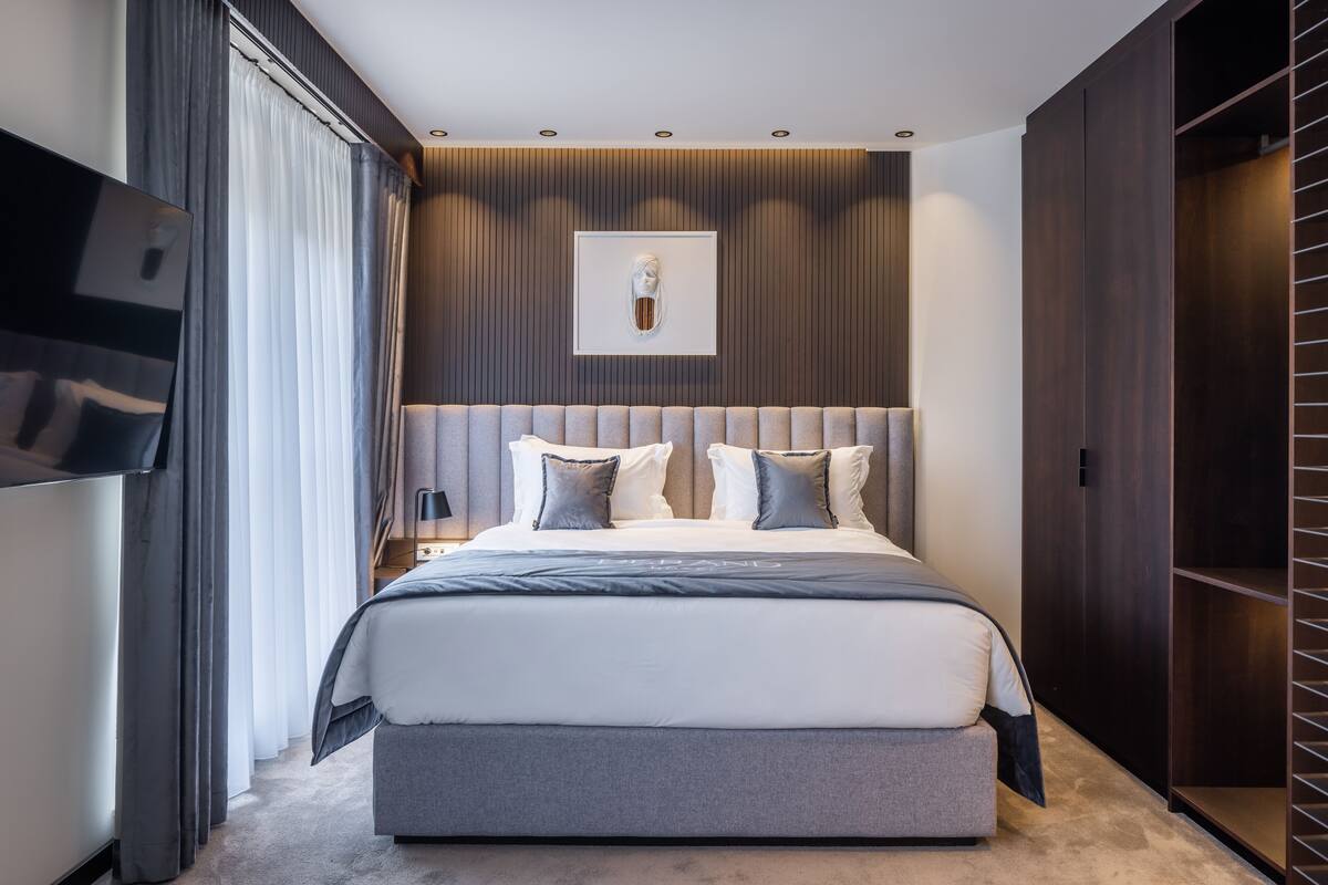 A modern bedroom with a large bed, a headboard with vertical lines, a framed artwork above the bed, and a wardrobe to the right.