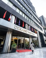 El exterior del Hotel Derand, con alfombra roja en la entrada, varias banderas y un diseño arquitectónico moderno.