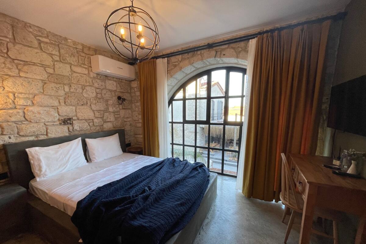 Un dormitorio con pared de piedra, cama con sábanas blancas, manta azul oscuro, escritorio de madera y una ventana arqueada grande con cortinas.