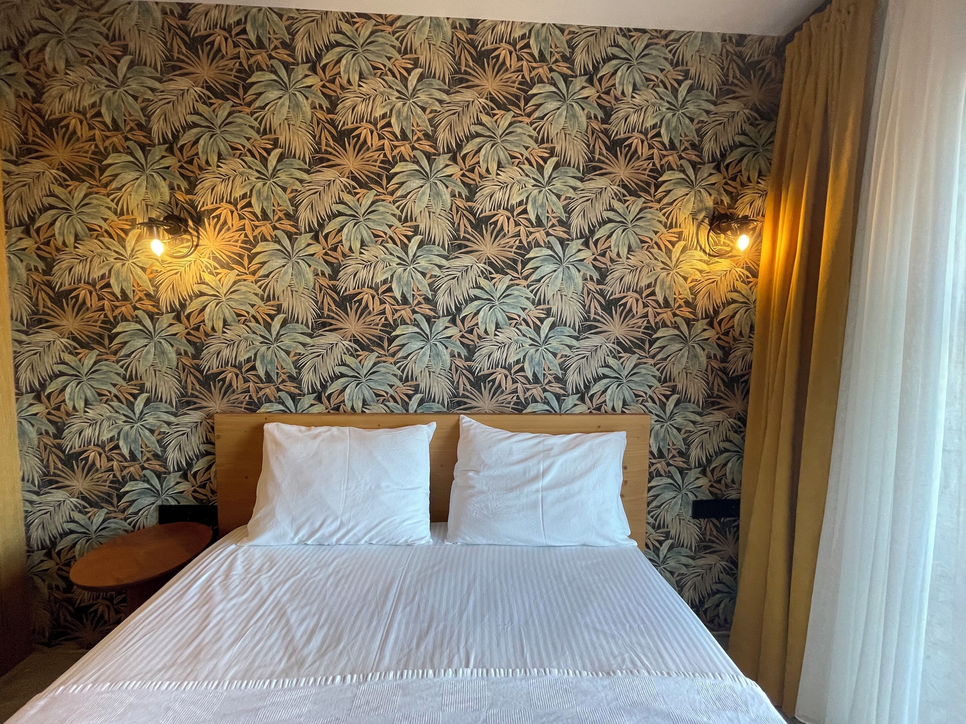 A hotel room with a bed, two pillows, a nightstand, and a wall with a tropical leaf pattern.