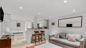 Un salon moderne avec un canapé, une table basse et des fauteuils. On aperçoit à l’arrière-plan un coin cuisine avec des placards et une table à manger.