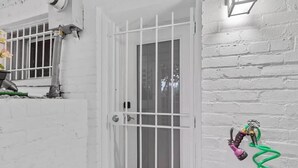 Une porte blanche avec une fenêtre grillagée, un tuyau d’arrosage vert et un luminaire fixé au mur.
