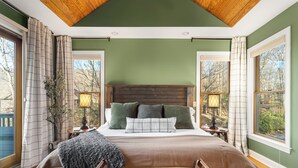 A bedroom with a large bed, wooden headboard, two bedside tables with lamps, and a view of trees through the windows. - Stunning Views, Hot Tub, Impeccable Design (Wintergreen Resort)