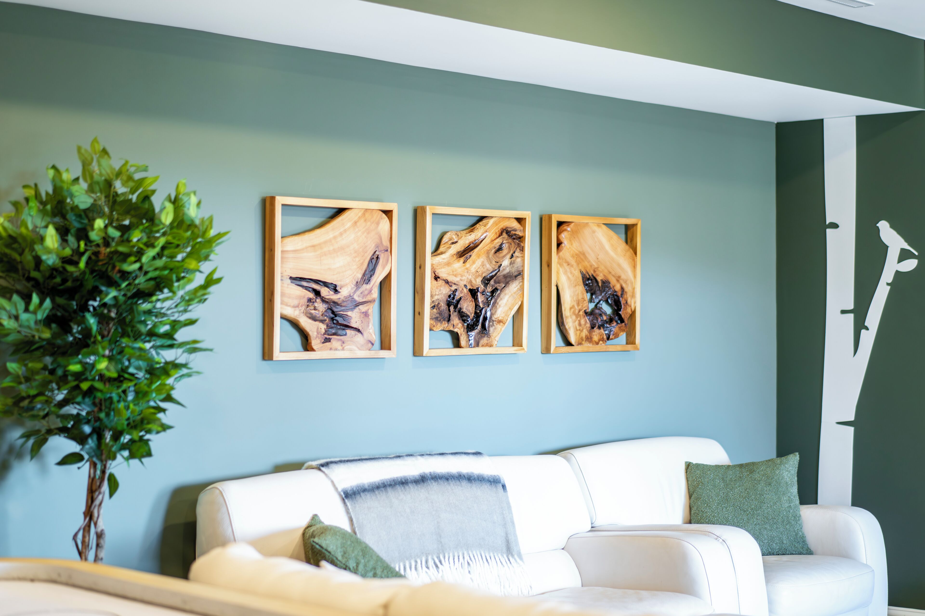 A modern living room with a white sofa, a green plant, and three abstract wooden art pieces on the wall.
