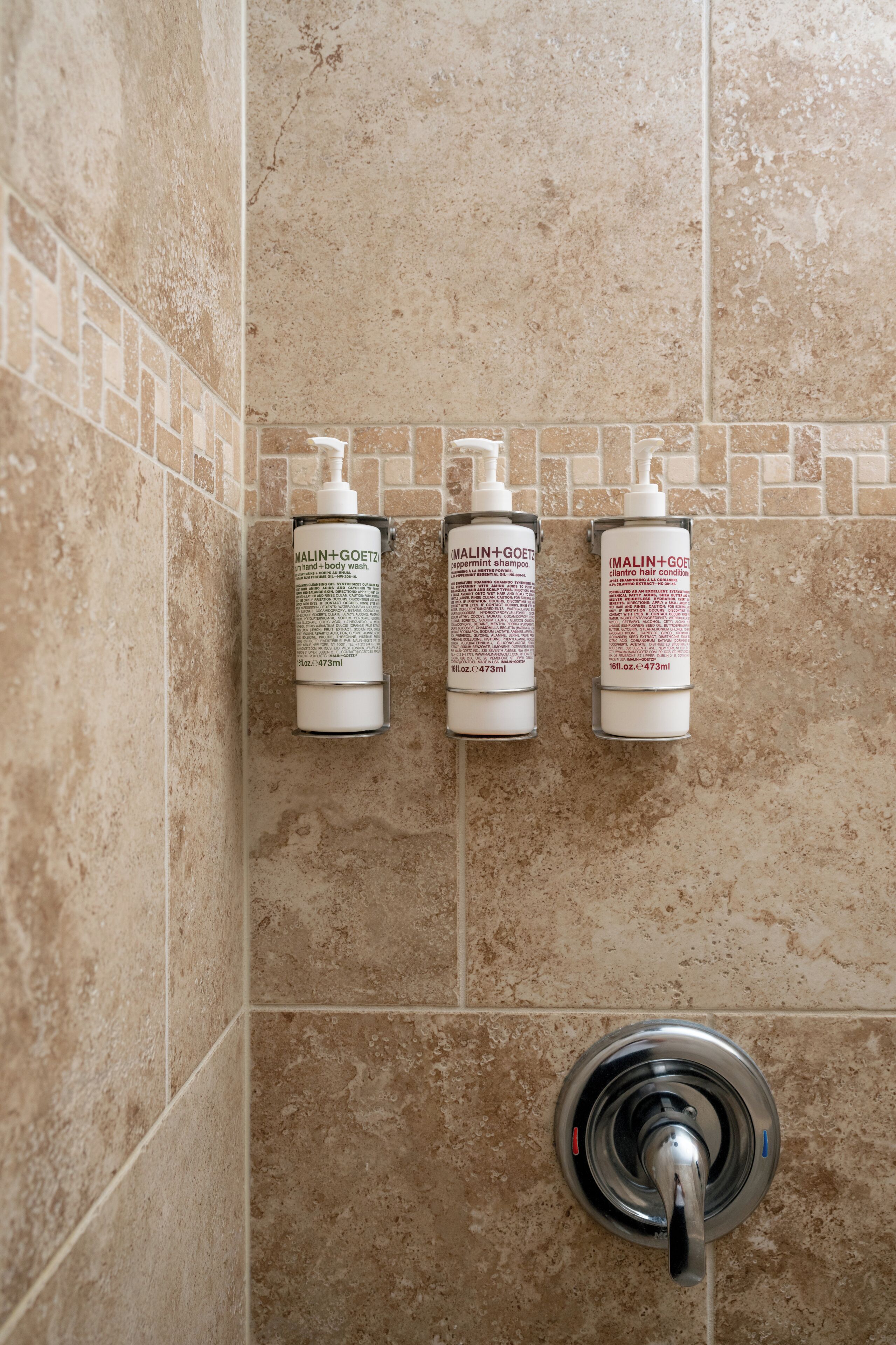 Three bottles of MALIN+GOETZ hand and body wash on a tiled wall.