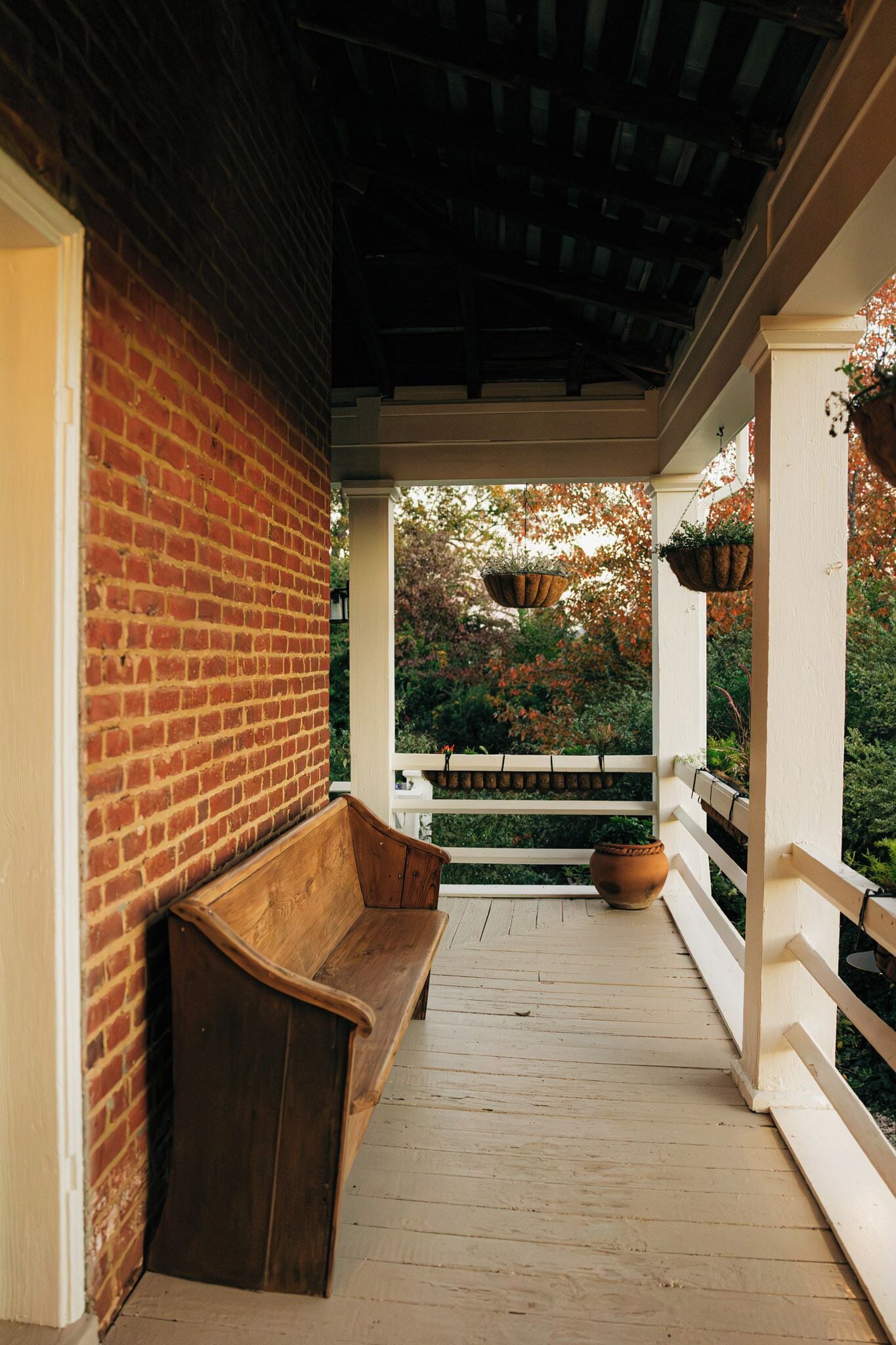 A covered porch with a wooden bench, a brick wall, and hanging planters.