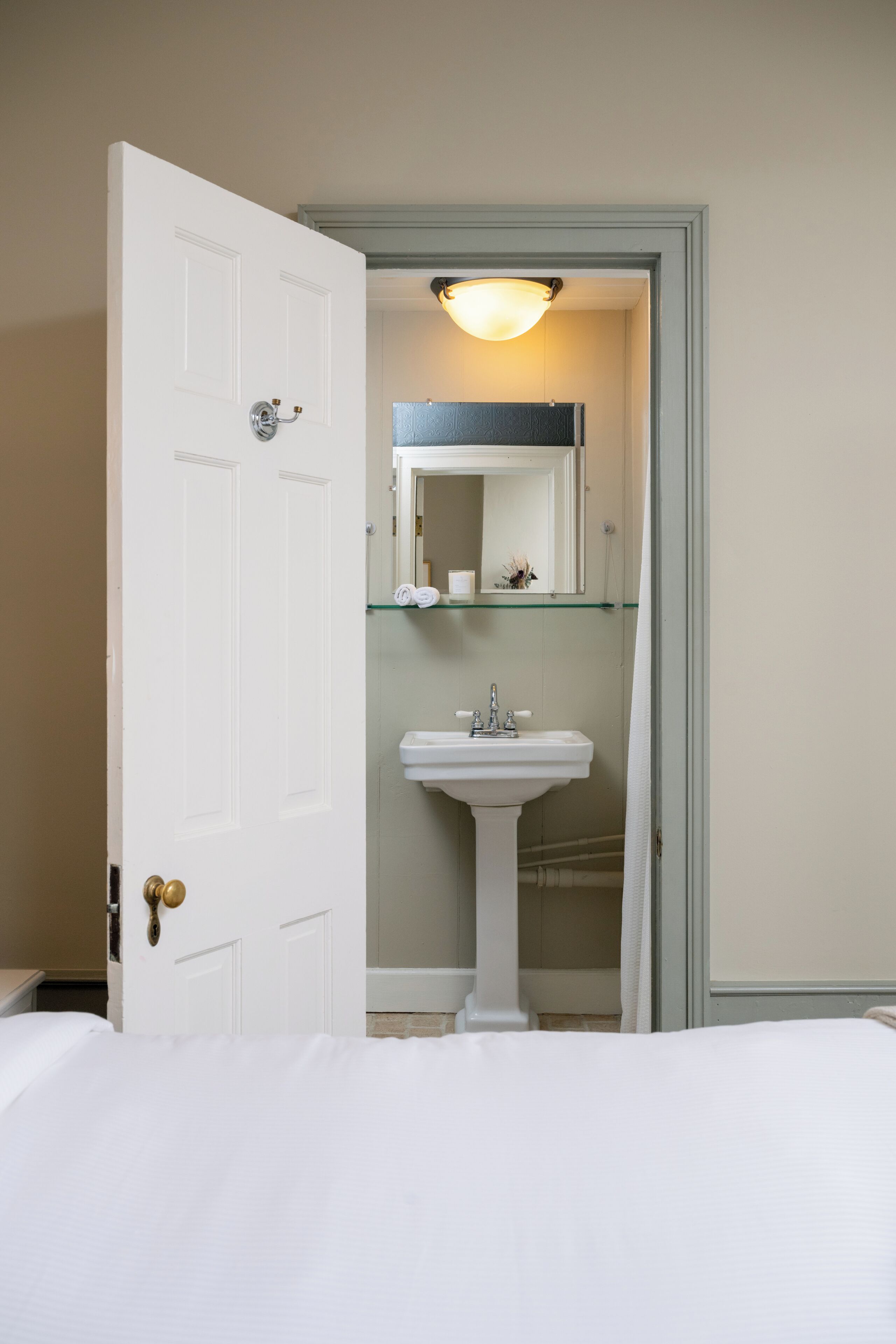 A hotel room with a white door, a bed, a white sink, and a glass shelf above the sink.