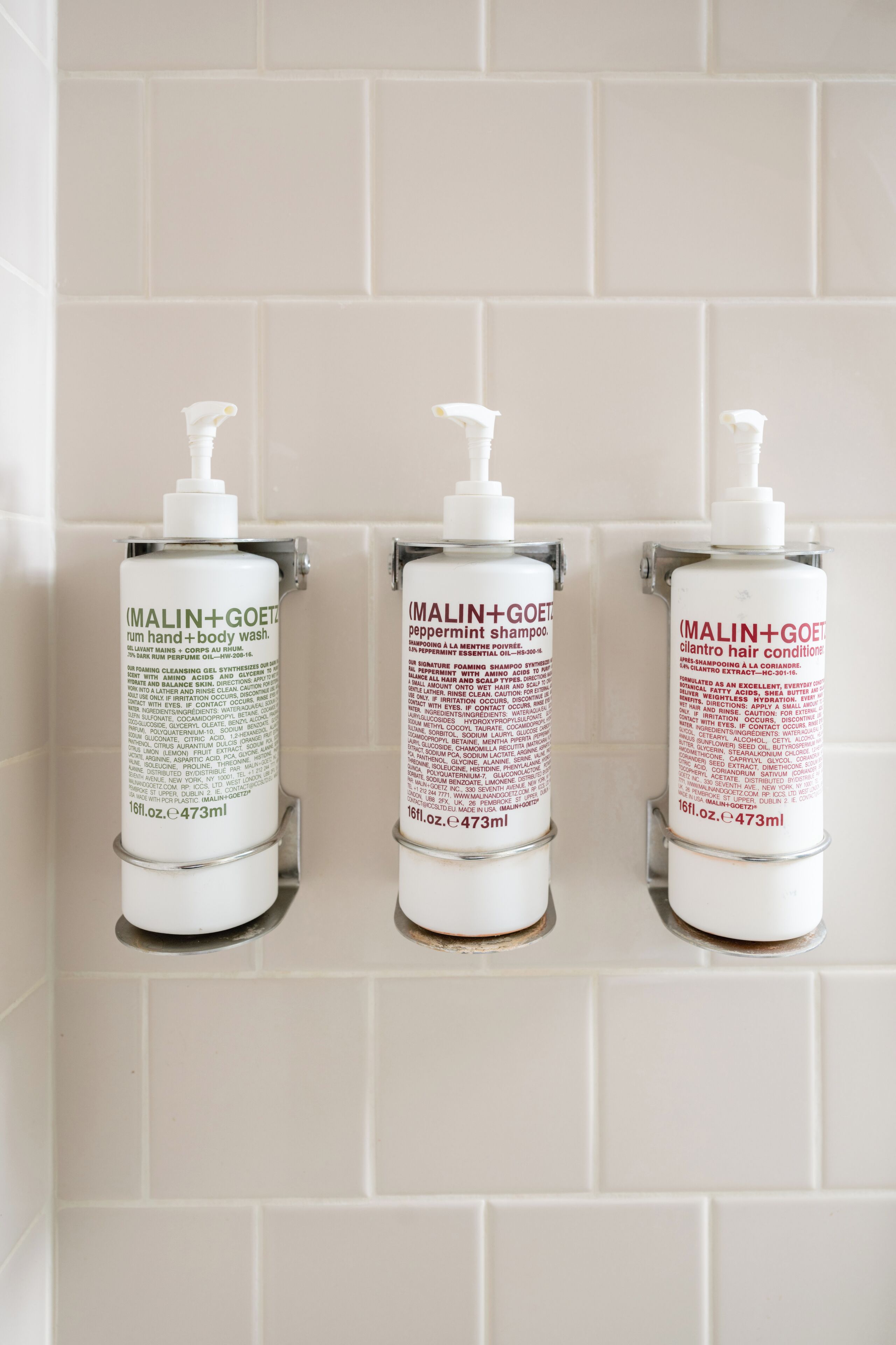 Three bottles of MALIN+GOET hand and body wash, and hair conditioner on a tiled wall.