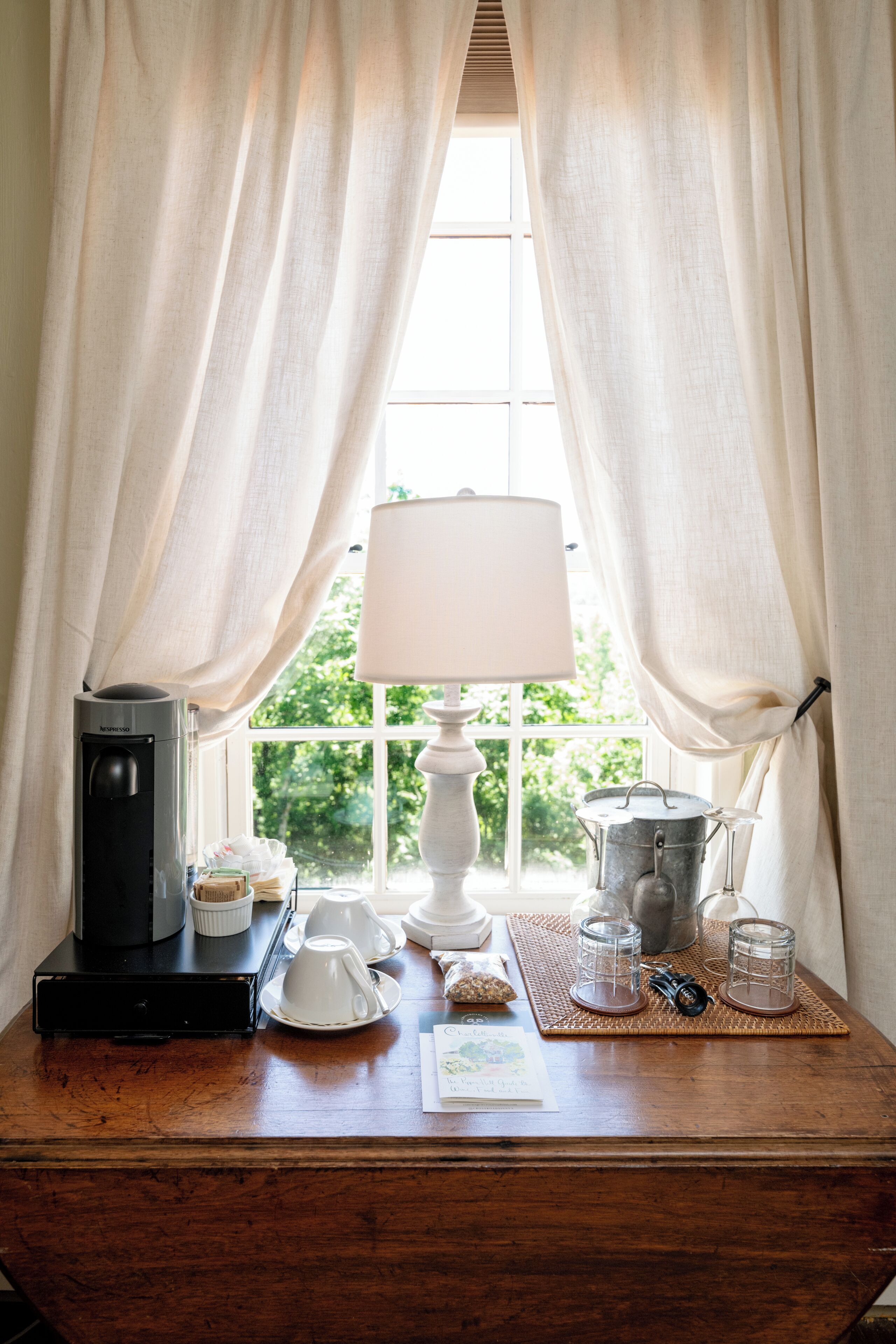 Ein Holztisch mit Kaffeemaschine, Teekanne, Tassen und einer Lampe in der Nähe eines Fensters mit Vorhängen.