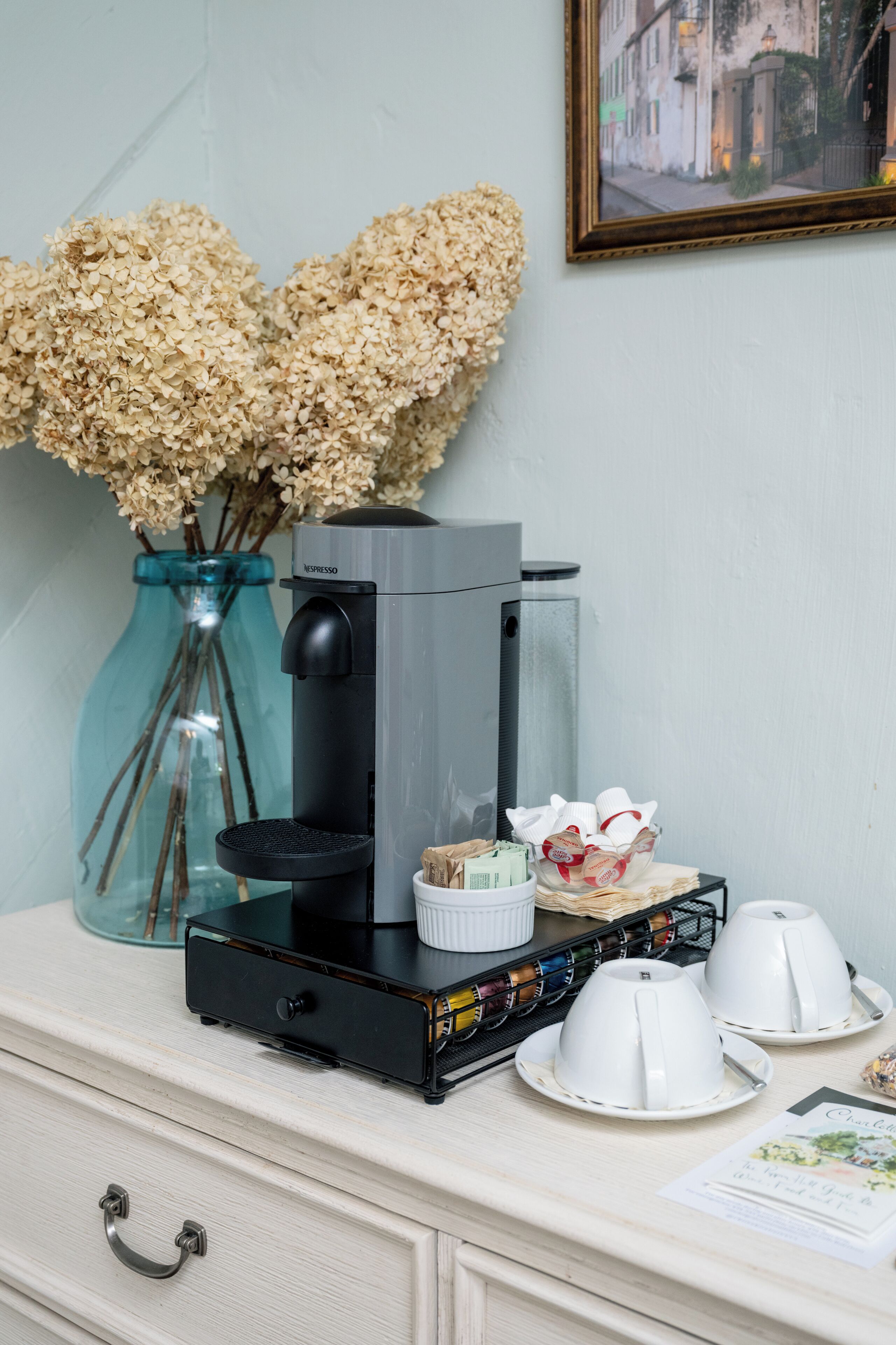 Une machine à café est posée sur un comptoir, à côté d’un vase rempli de fleurs séchées et d’un tableau encadré accroché au mur.