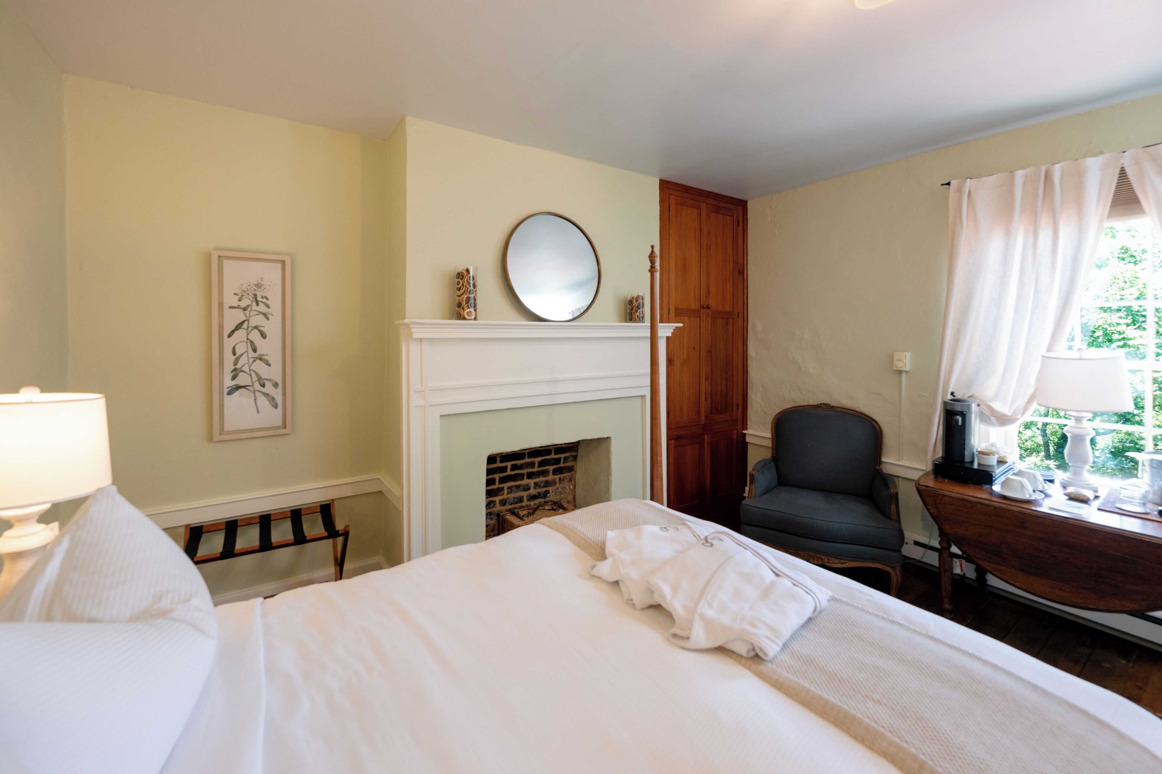 A bedroom with a bed, a chair, a desk, a fireplace, and a window with curtains.