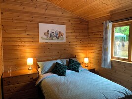 A wooden cabin room with a bed, two bedside lamps, a window with curtains, and a painting of animals on the wall.