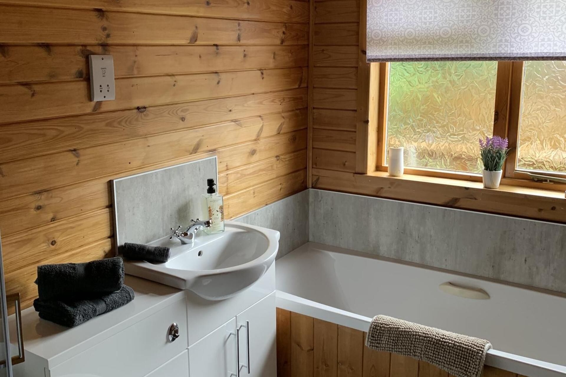 A wooden cabin bathroom with a white sink, a mirror, and a window with a view of greenery.