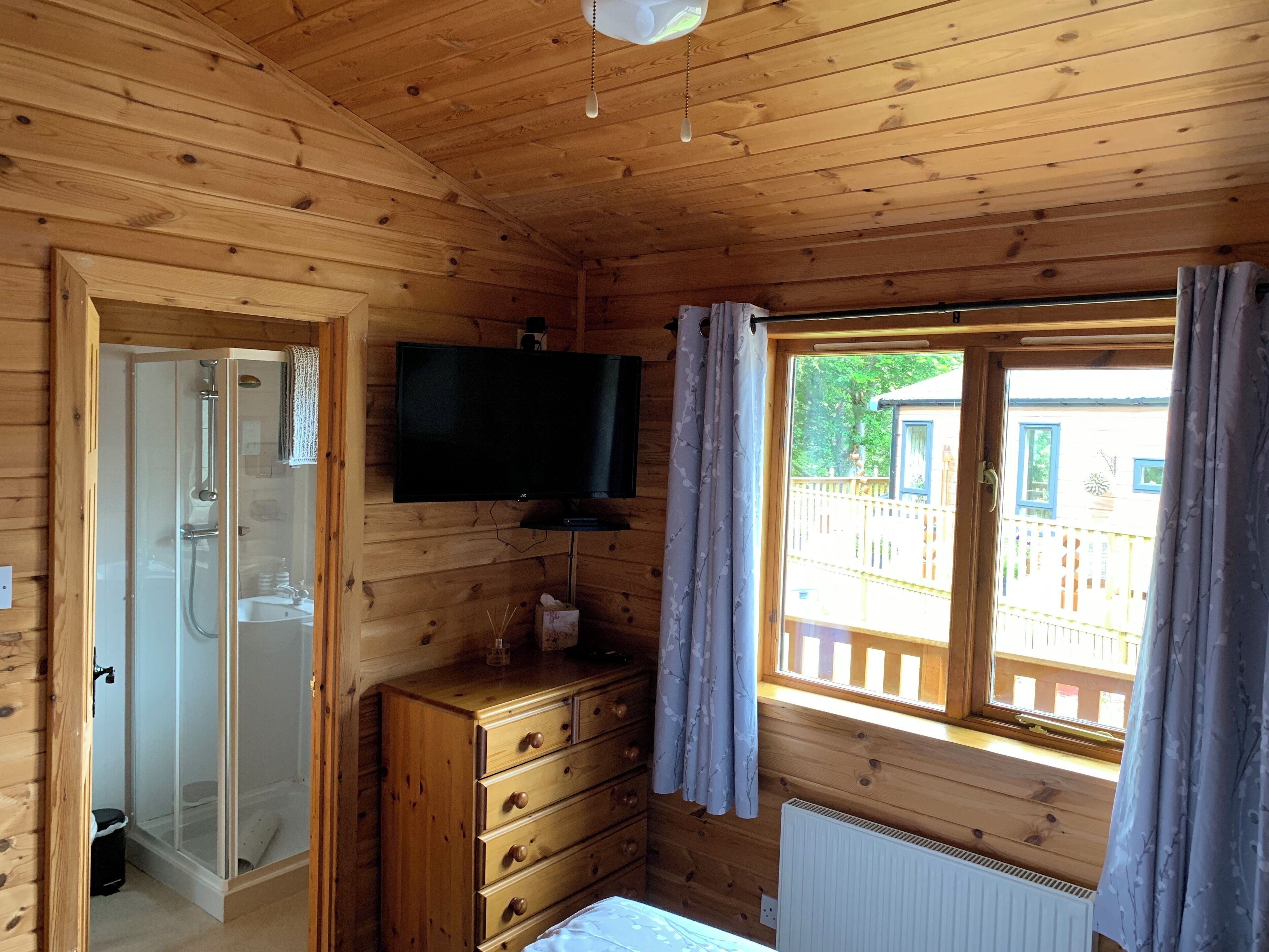 Une pièce de style cabane en bois, équipée d’une douche, d’une commode en bois, d’un téléviseur fixé au mur et d’une fenêtre avec des rideaux.