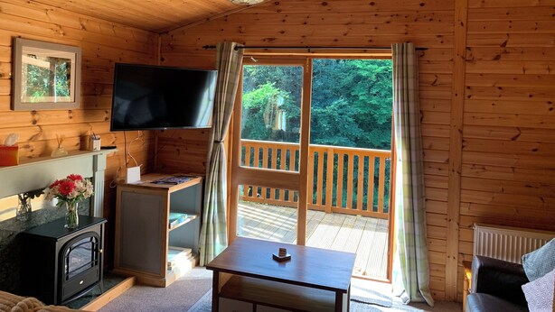 A cozy cabin interior with a wooden ceiling, a television mounted on the wall, a small table, and a window with curtains. - Lodge With River View - Sleeps 6 - pet Friendly (Hawick)