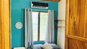 A small, cozy room with a bed, a window with curtains, a small table, and a wall-mounted air conditioner.