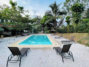 A rectangular swimming pool with a tiled edge, surrounded by a gravel area. There are four black lounge chairs aligned in a row facing the pool. - Apartamentos El Labrador # 7 (Tamarindo)