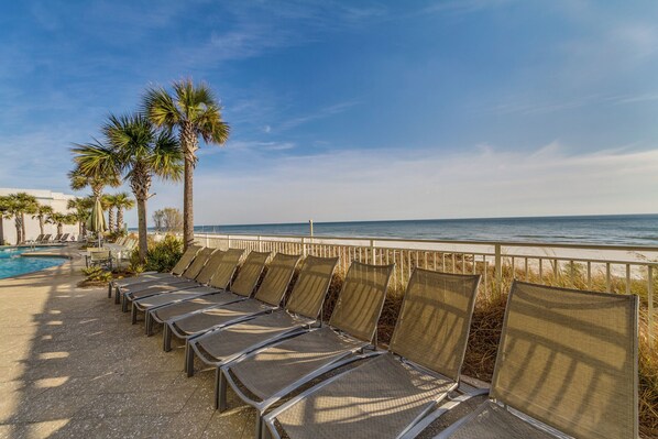A poolside area with lounge chairs and palm trees, overlooking a beach and ocean.