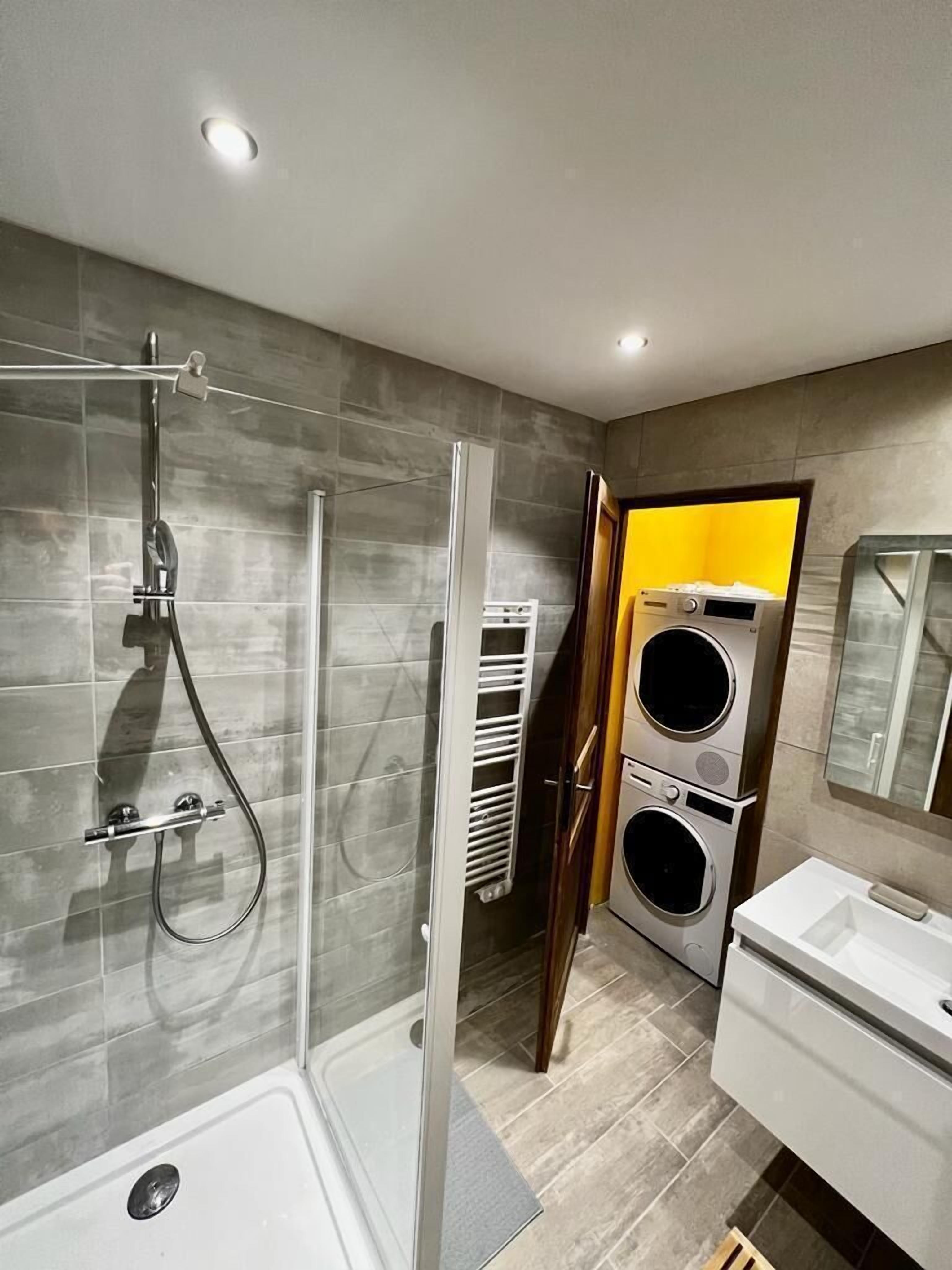 A modern bathroom with a glass shower enclosure, a built-in washing machine, and a vanity with a mirror.