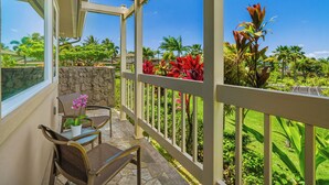 A balcony with a stone wall, a table with chairs, and a view of a lush garden with palm trees and red foliage.