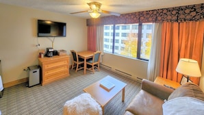 Une chambre d’hôtel équipée d’un téléviseur à écran plat, d’un ventilateur de plafond, d’une table à manger avec des chaises, d’un canapé, d’une petite table avec une lampe et d’une fenêtre avec des rideaux.