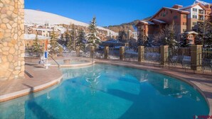 Un espace piscine agrémenté d’un mur en pierre, d’un jacuzzi et offrant une vue sur des bâtiments et des arbres recouverts de neige.