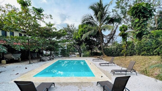 A rectangular swimming pool with a tiled edge, surrounded by a gravel area. There are four black lounge chairs aligned in a row facing the pool. - Apartamentos El Labrador # 10 (Tamarindo)