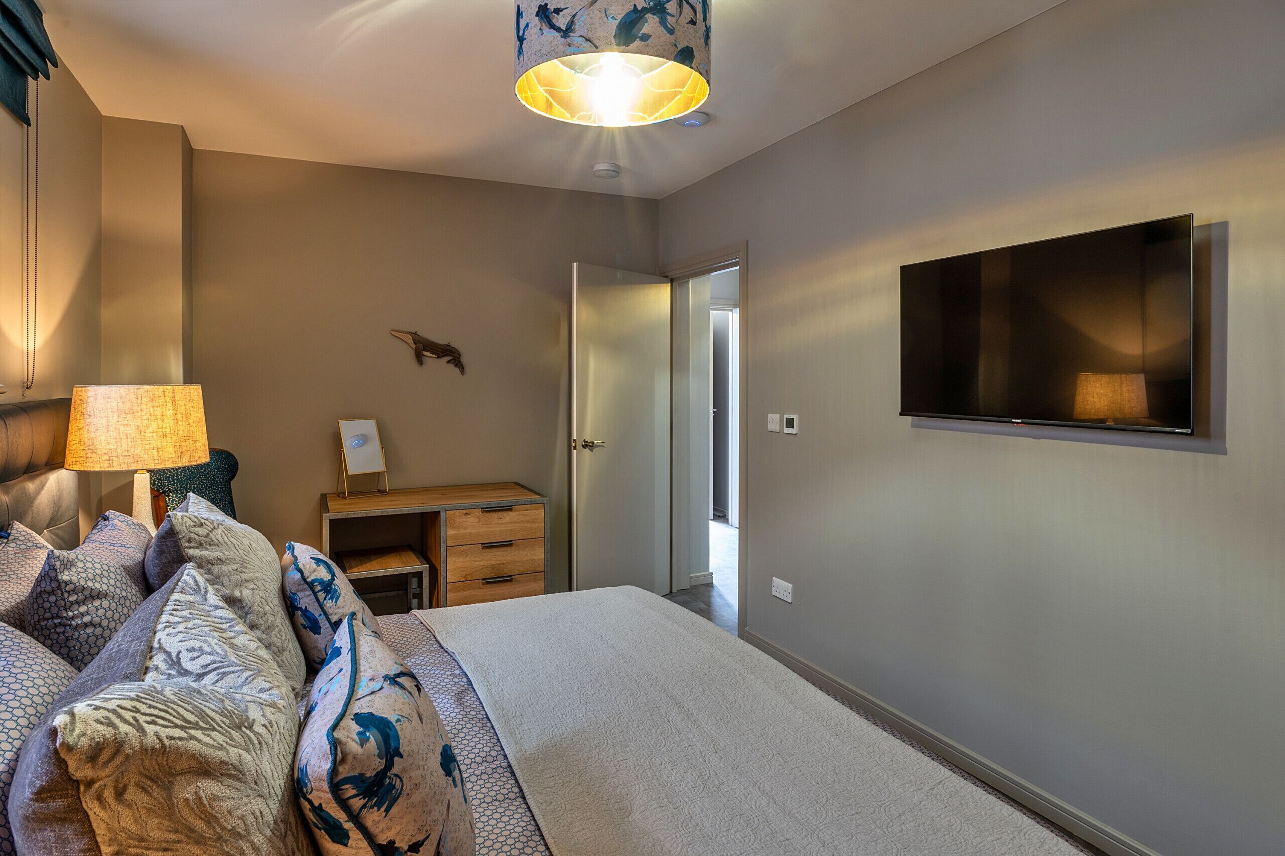 A bedroom with a bed, a desk, a TV, and a door leading to another room.