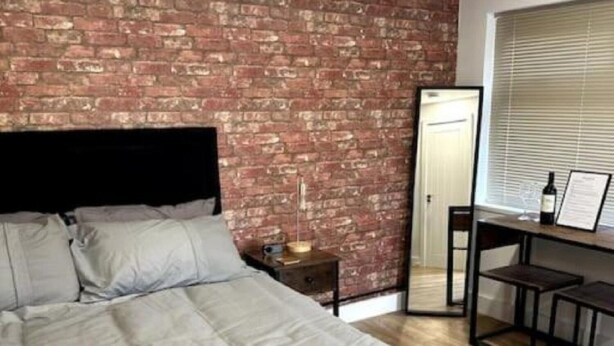A modern bedroom with a brick wall, a bed with grey bedding, a nightstand, a desk with a chair, and a window with blinds. - Perfect stay with Dallas Studio (Hayes)