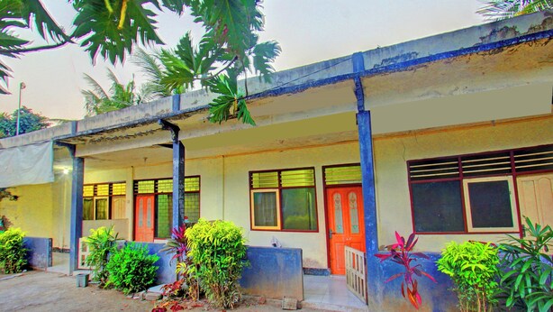 A building with a covered porch, multiple windows, and a door. There are plants and a concrete bench in front. - Hotel O Penginapan 3 Bersaudara Near Pasar Sengkol (Janapria)