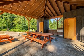 Enjoy dining and socializing in this outdoor patio area with a wooden table and benches, sheltered by a roof structure.