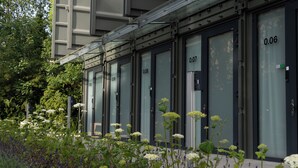 Una fila de habitaciones con fachadas de vidrio, números visibles y un pequeño jardín exterior con flores.