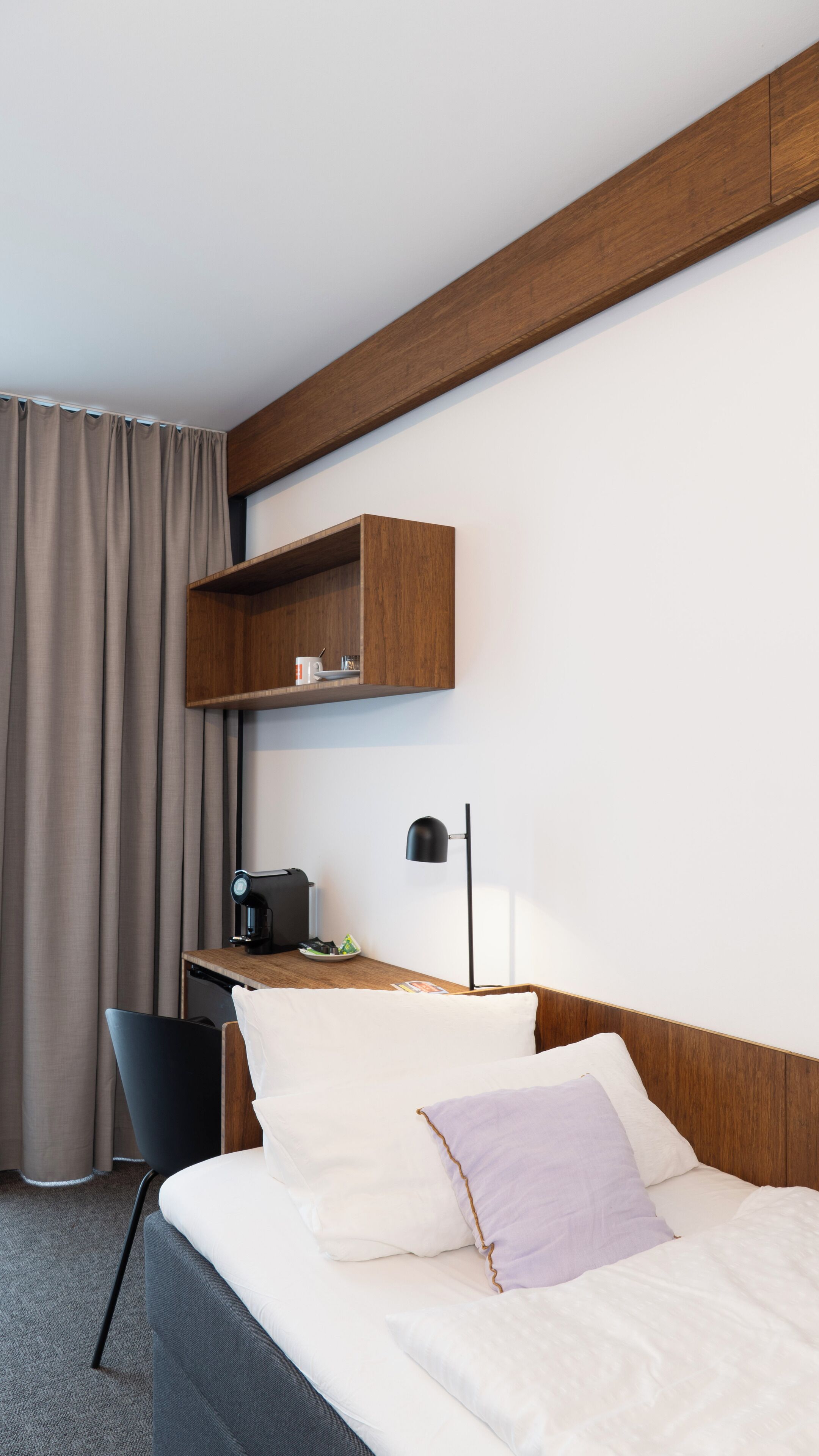 A hotel room with a bed, a desk with a lamp, a chair, and a shelf above the desk.