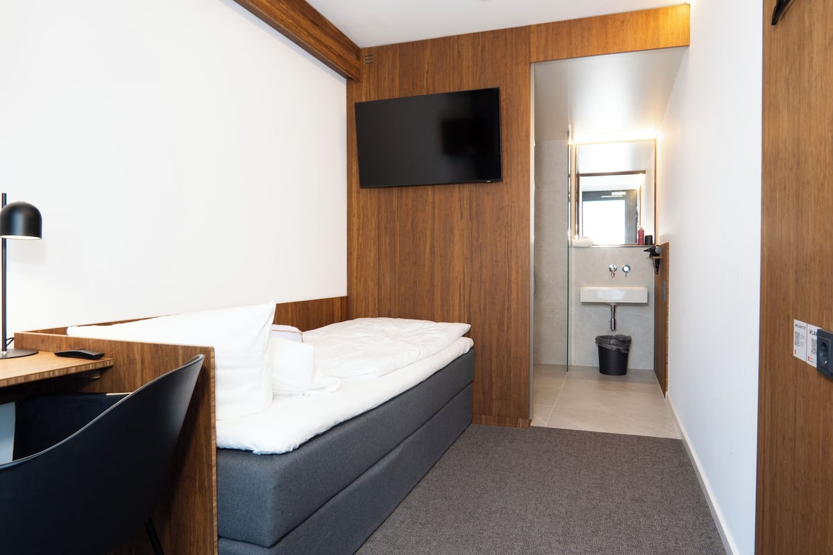 A hotel room with a bed, desk, chair, TV, and a small bathroom with a sink and mirror.